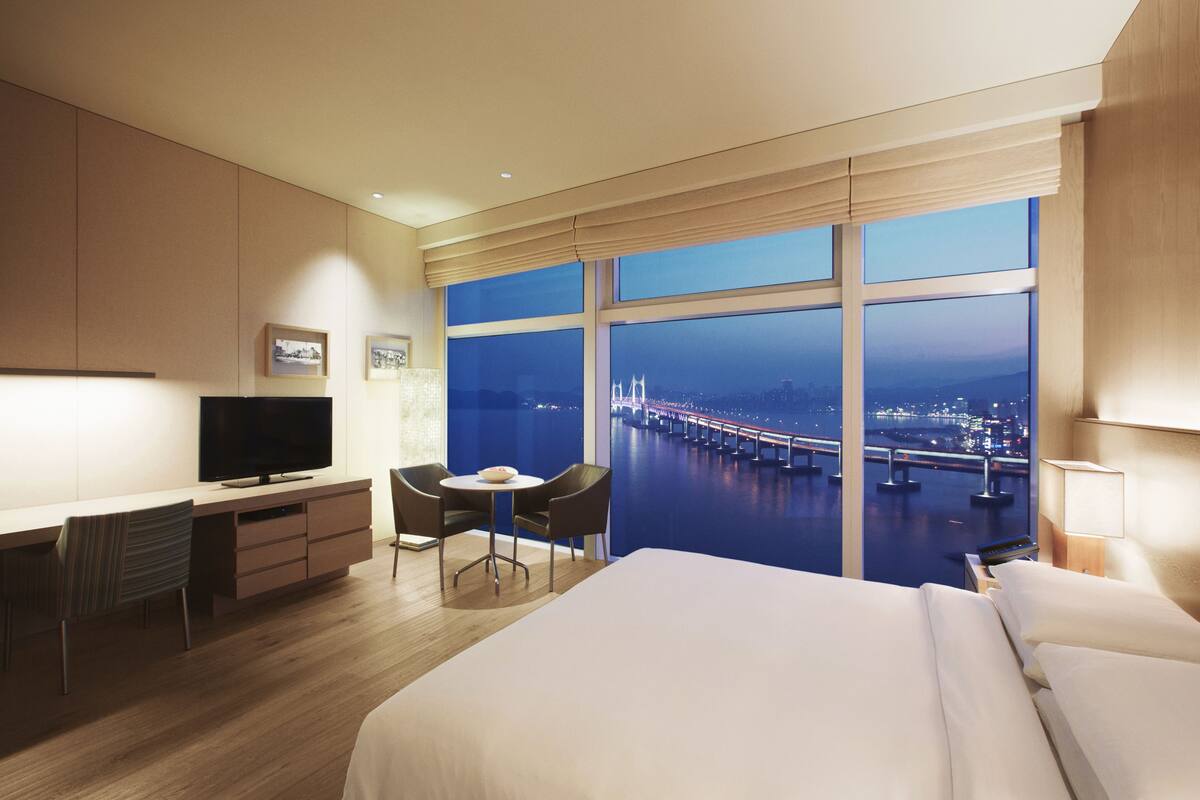 A hotel room with a large bed, a desk with a TV, a small table with chairs, and a view of a cityscape and bridge at night.