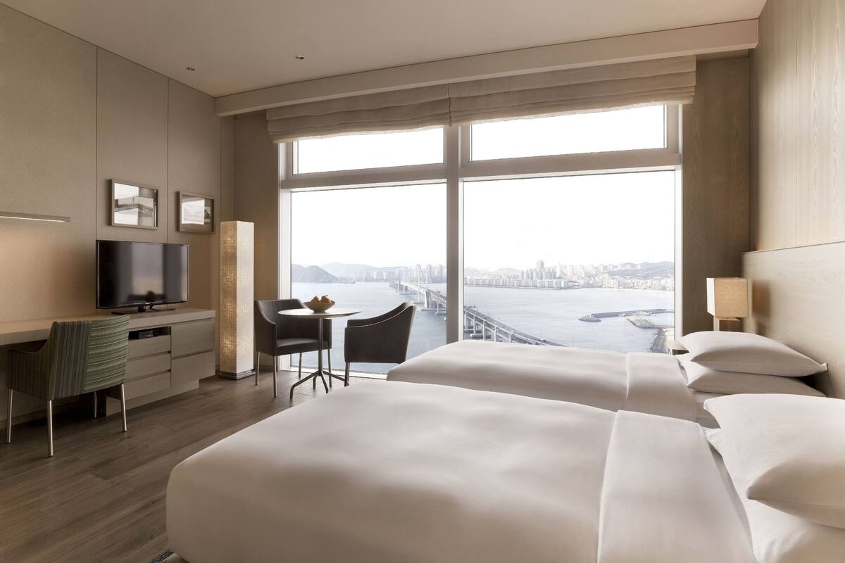 A hotel room with two beds, a desk with a TV, a chair, and a view of a bridge and cityscape.