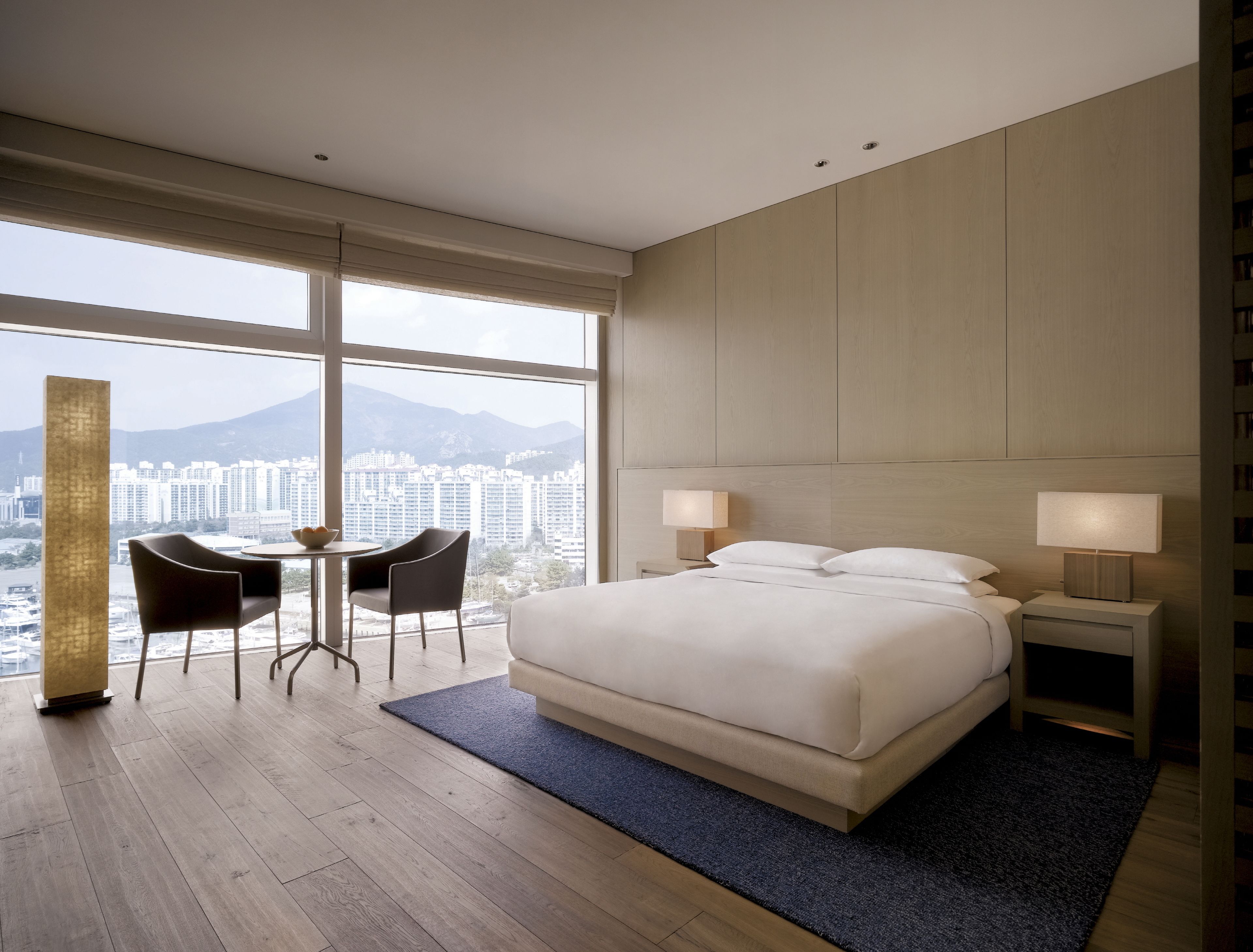 Un hotel moderno con una cama grande, dos mesitas de noche con lámparas, una pequeña mesa con sillas y vistas a un paisaje urbano a través de amplias ventanas.