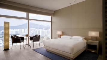 Un hotel moderno con una cama grande, dos mesitas de noche con lámparas, una pequeña mesa con sillas y vistas a un paisaje urbano a través de amplias ventanas.