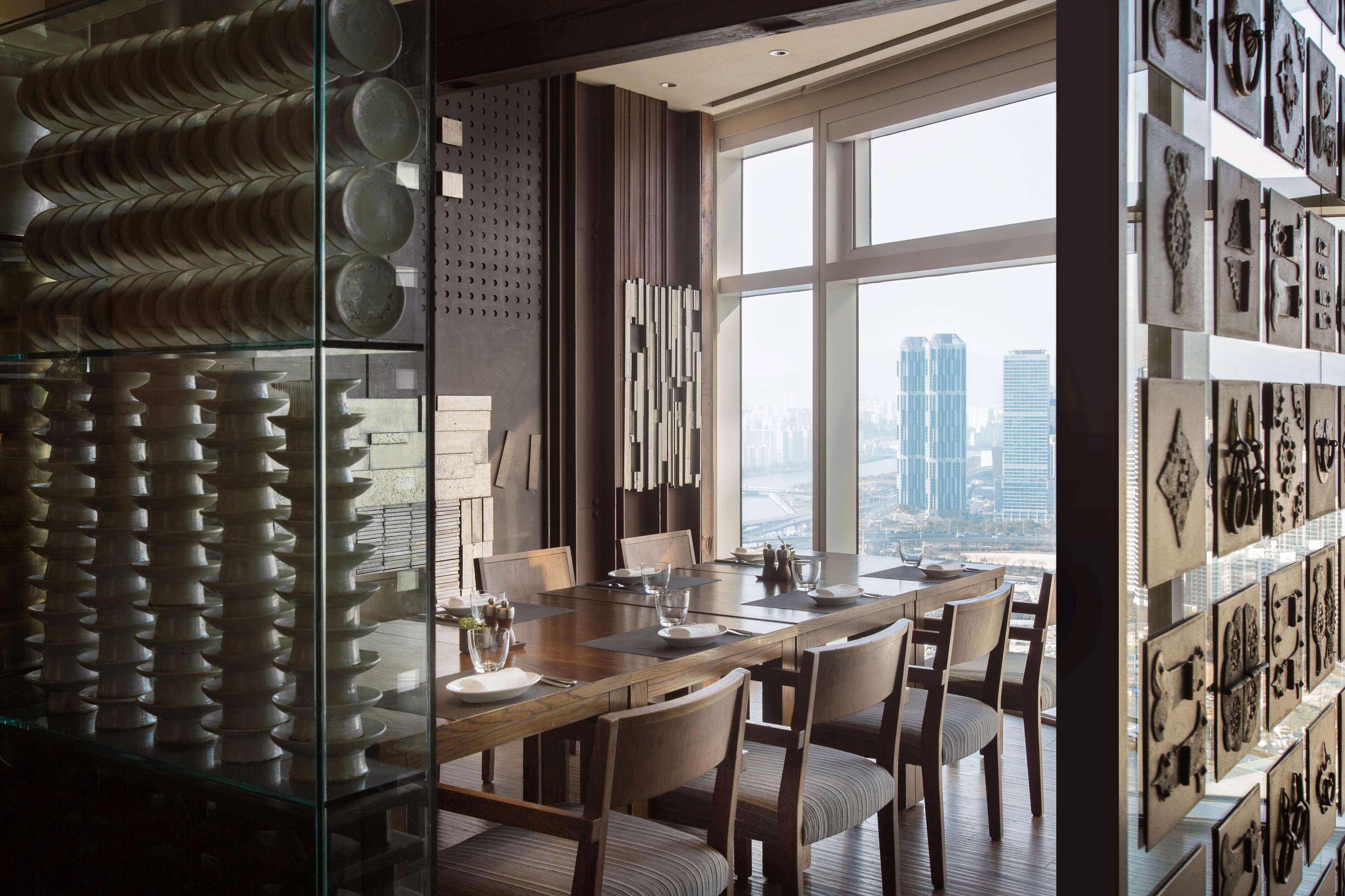Un comedor moderno con una mesa grande y sillas, vista a la ciudad a través de amplios ventanales, y una pared decorada con varios objetos.