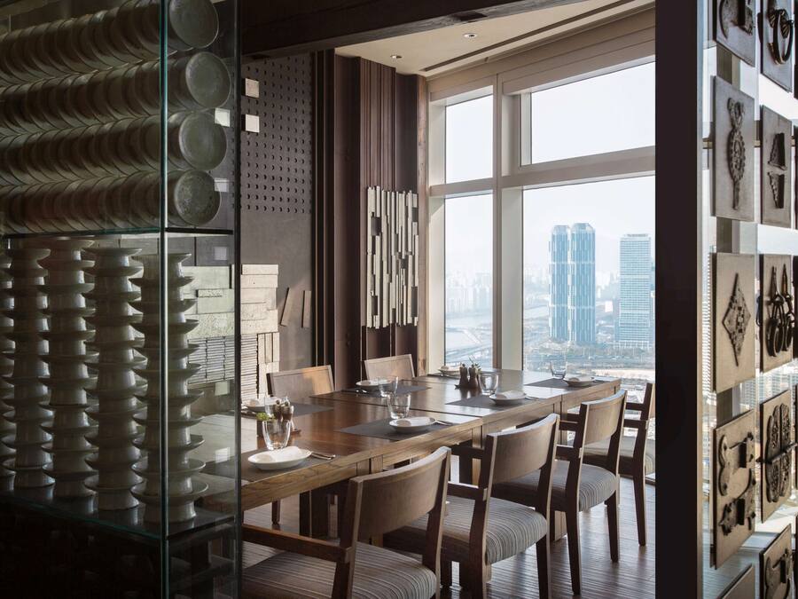 Un comedor moderno con una mesa grande y sillas, vista a la ciudad a través de amplios ventanales, y una pared decorada con varios objetos.