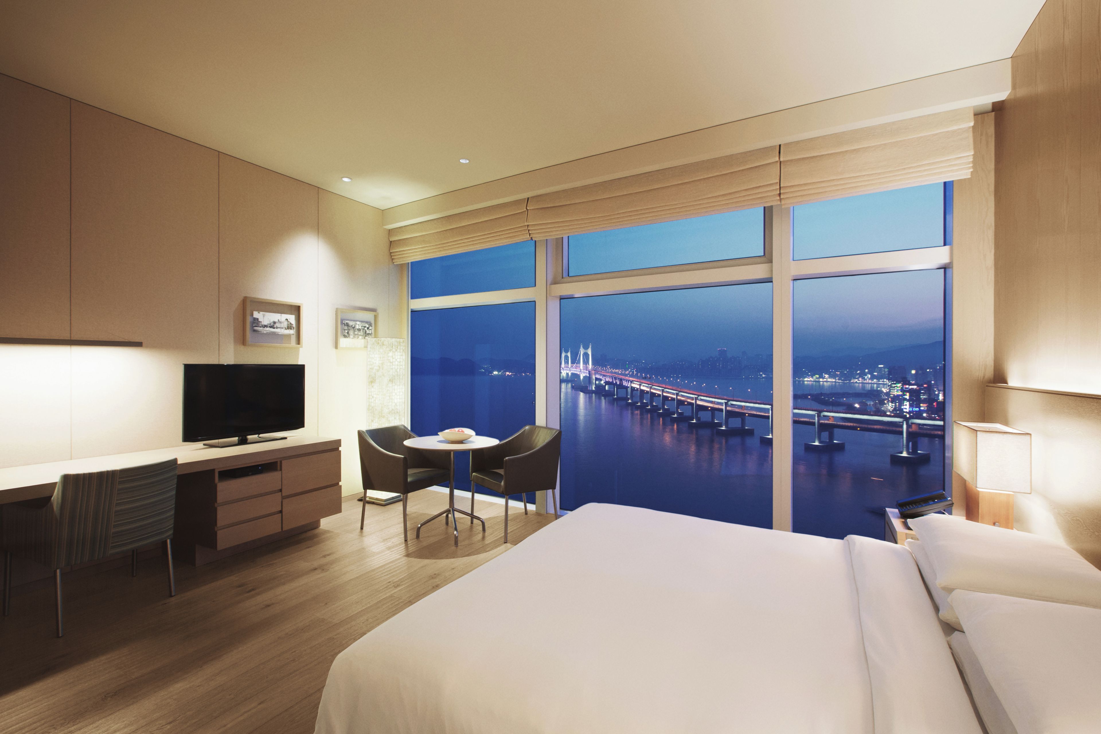 Habitación de hotel con una cama, un escritorio con televisor, una silla, una mesita y vista a un puente de noche.