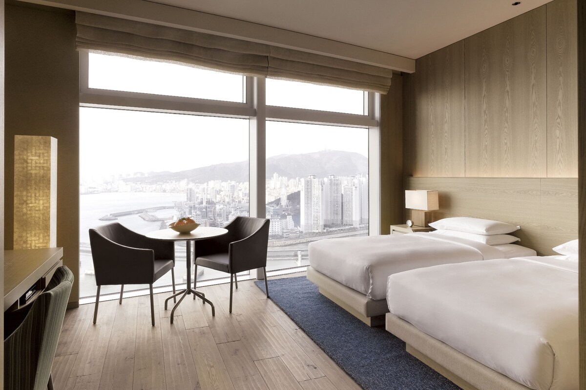 A hotel room with a large window, two beds, a desk with chairs, and a view of a cityscape.