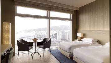 Habitación de hotel con un ventanal grande, dos camas, un escritorio con sillas y vistas a la ciudad.