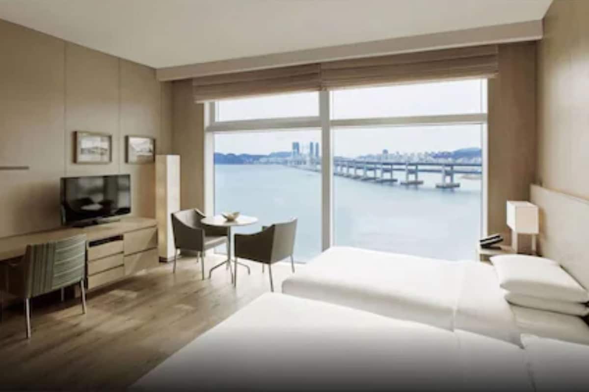 A hotel room with a bed, a desk with a chair, a television, and a large window with a view of a river and bridge.