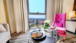 Quarto de hotel com uma janela ampla, uma cadeira rosa, uma mesa com petiscos e vista para a paisagem urbana.