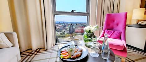 A hotel room with a large window, a pink chair, a table with snacks, and a view of a cityscape.
