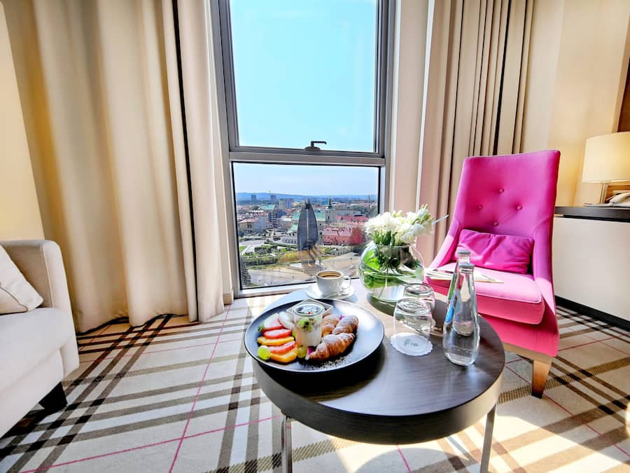 Una habitación de hotel con un ventanal grande, una silla rosa, una mesa con aperitivos y vistas a un paisaje urbano.