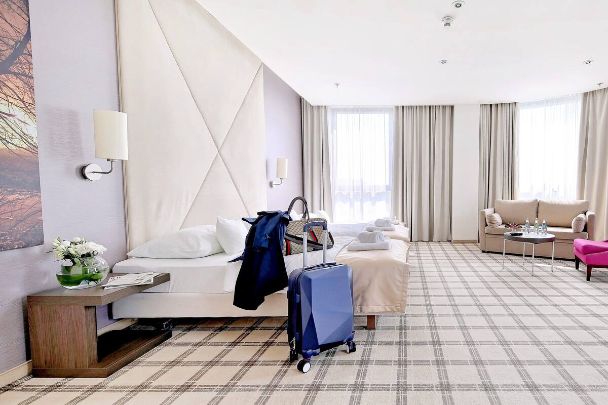 A hotel room with a bed, a sofa, a pink chair, a blue suitcase, and a table with bottles.