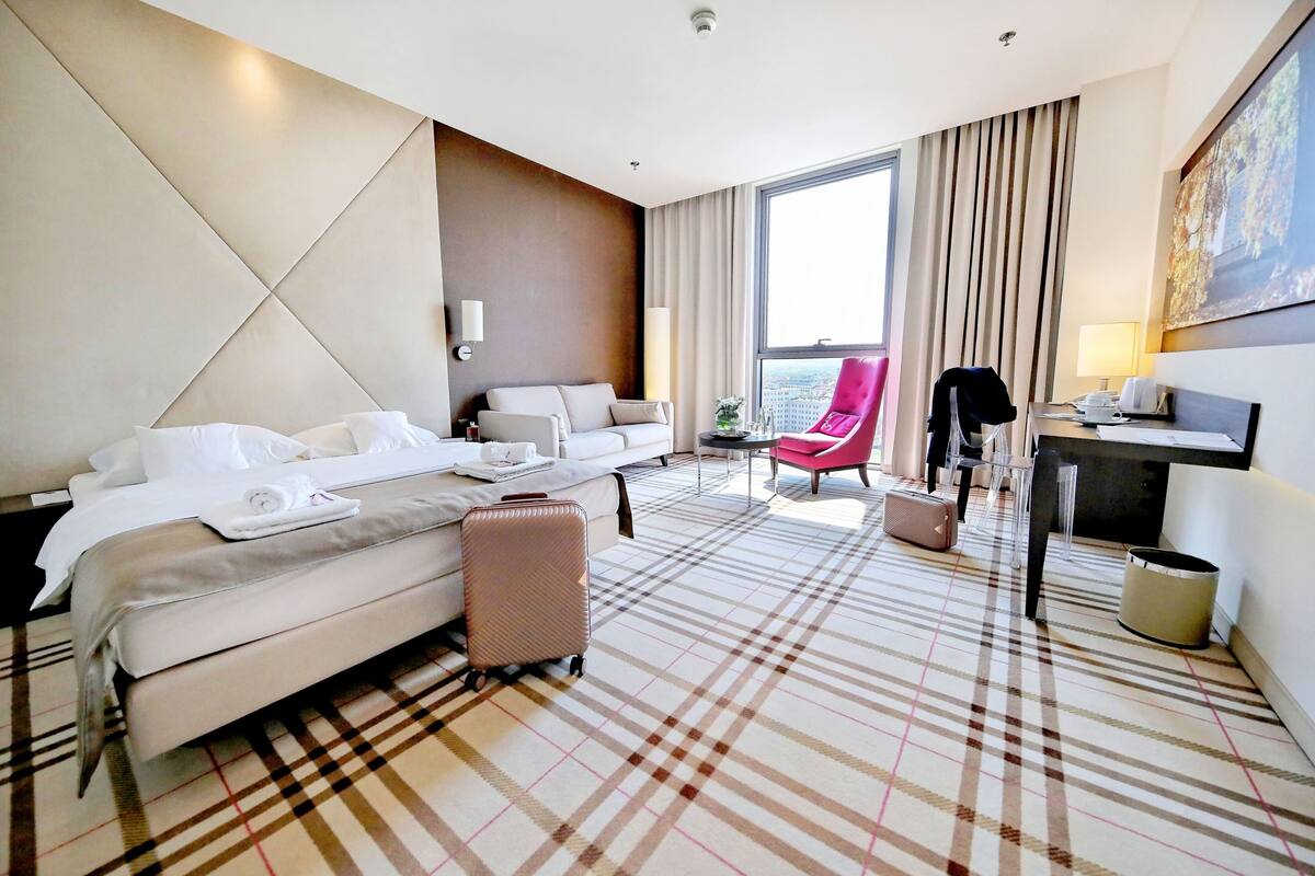 A hotel room with a large bed, a sofa, a red armchair, a desk with a lamp, and a view of the city.