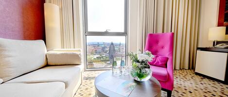 Habitación de hotel con un ventanal grande, un sillón rosa, un sofá blanco, una mesa redonda con un jarrón de flores y un mostrador de recepción.