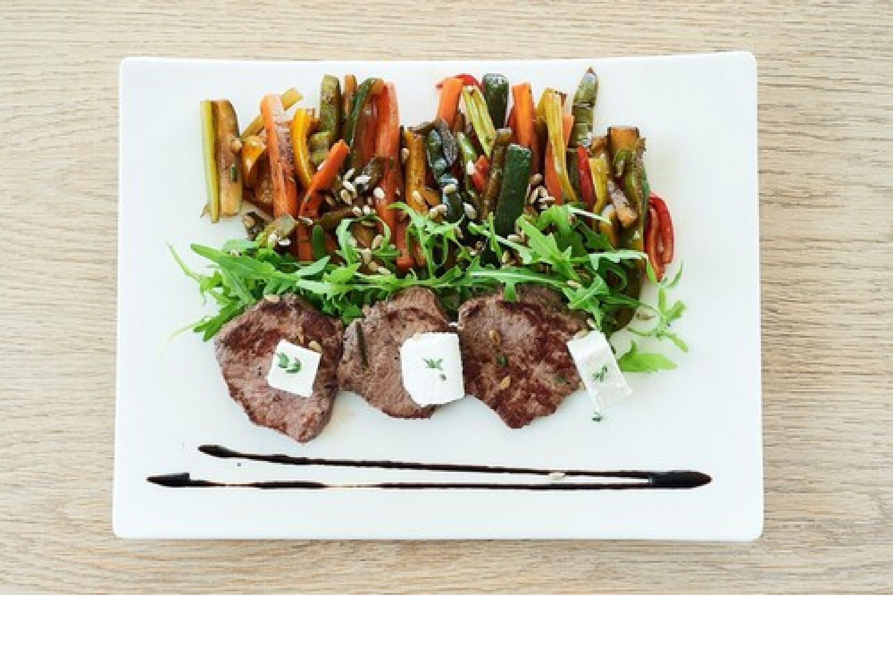 A white plate with a variety of vegetables and three pieces of meat on a wooden table.