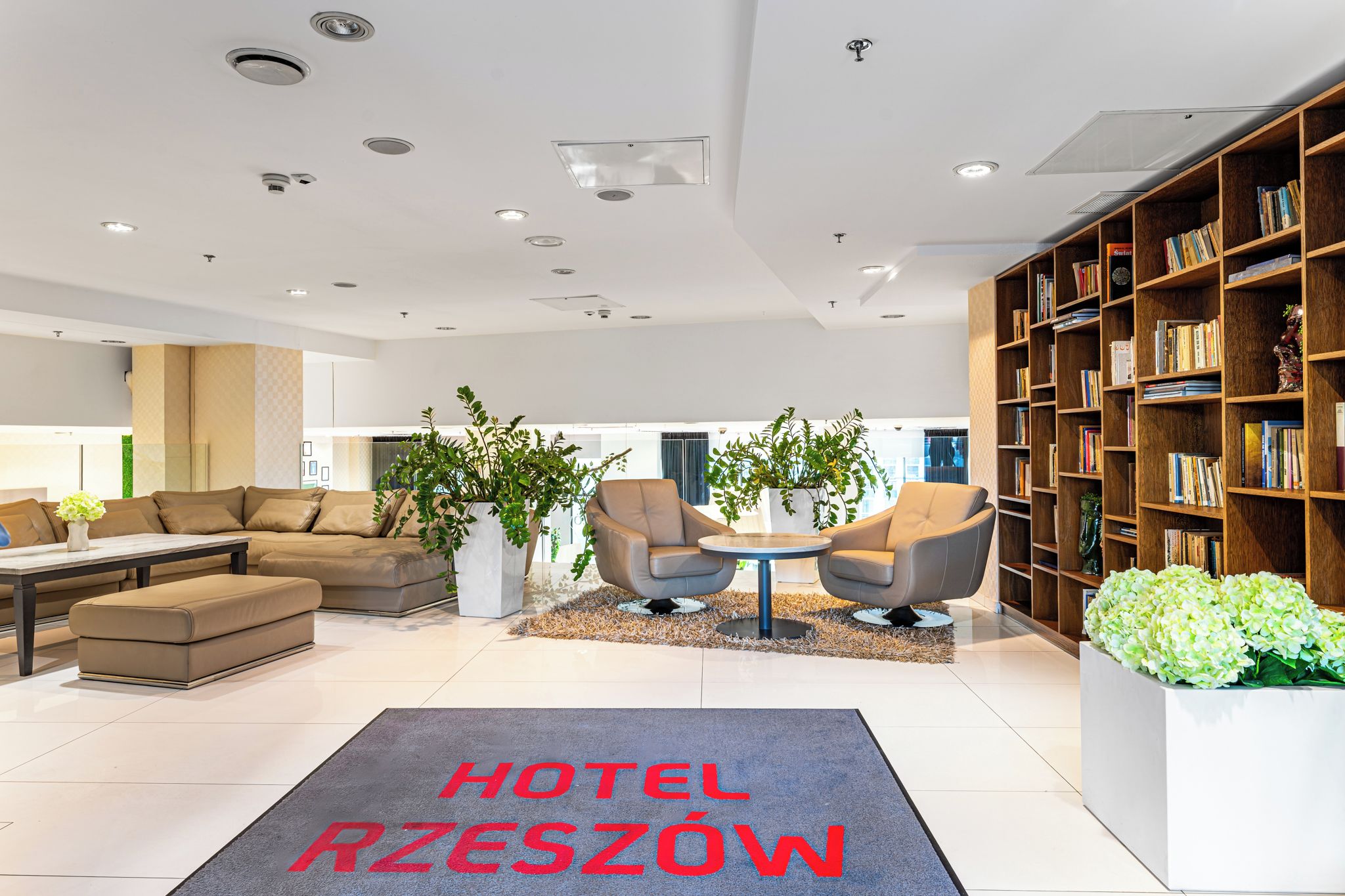 Un moderno vestíbulo de hotel con un conjunto de sofás, dos sillones, una mesa de centro, una estantería y un mostrador de recepción.