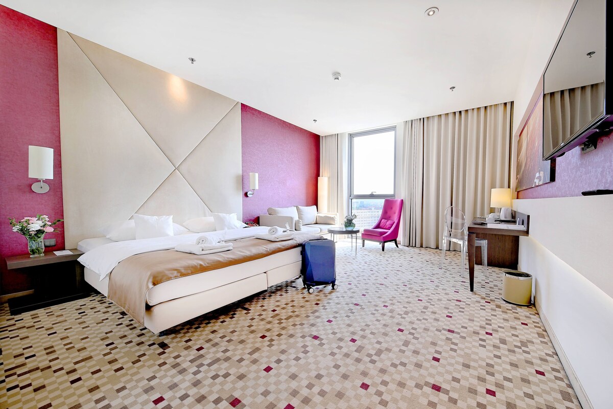 A hotel room with a large bed, a desk, a chair, and a view of the city through the window.