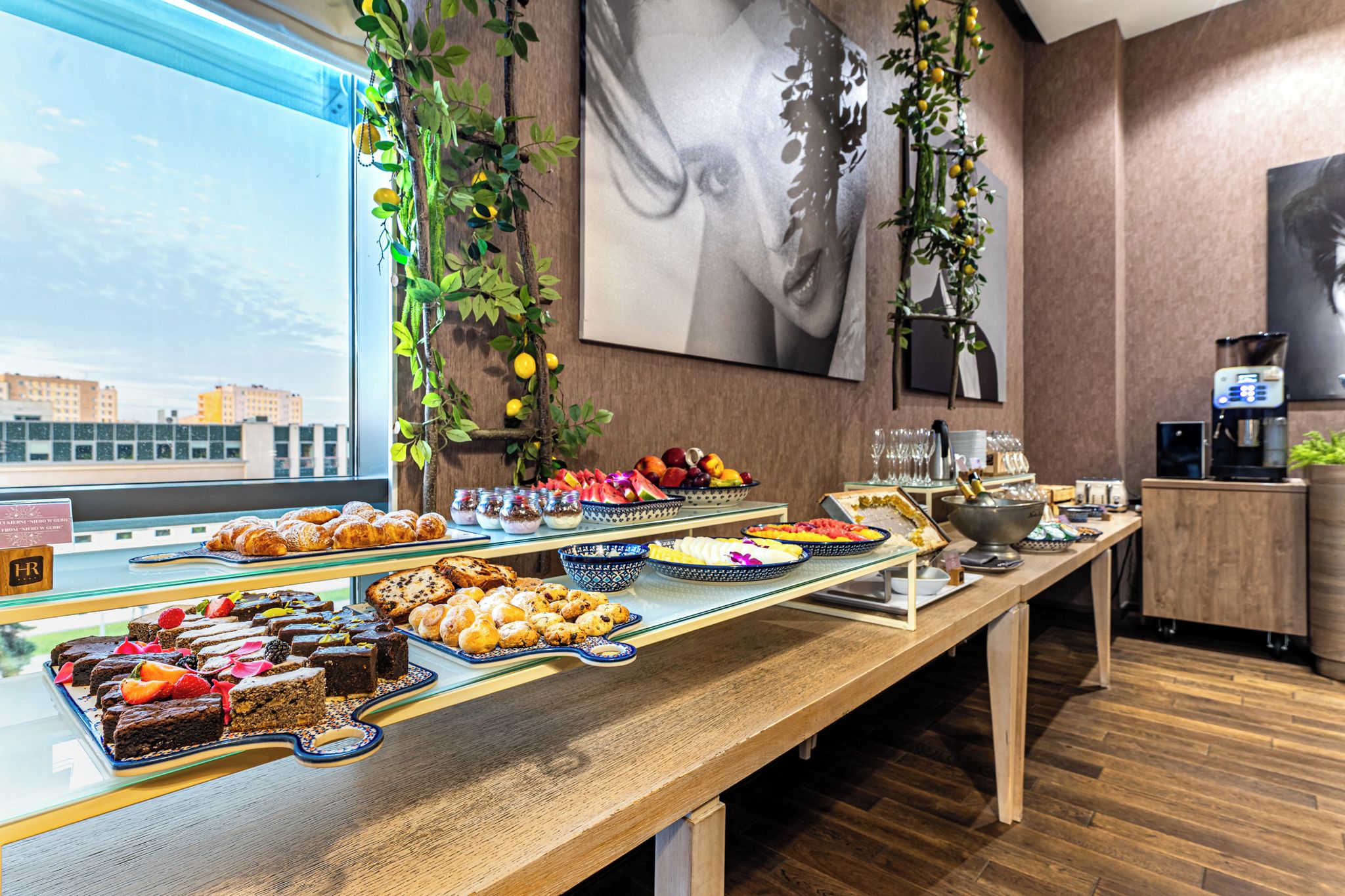 Un bufé con diversos postres y frutas, una máquina de café y vistas de la ciudad a través de una ventana.