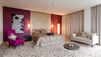 Una habitación de hotel con una cama, un sillón rosa, un sofá blanco, una mesa de centro redonda y un suelo estampado.