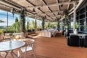 An indoor dining area with tables and chairs, large windows, and hanging plants.