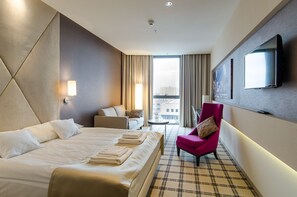 A hotel room with a large bed, a small sofa, a chair, a TV, and a window with curtains.