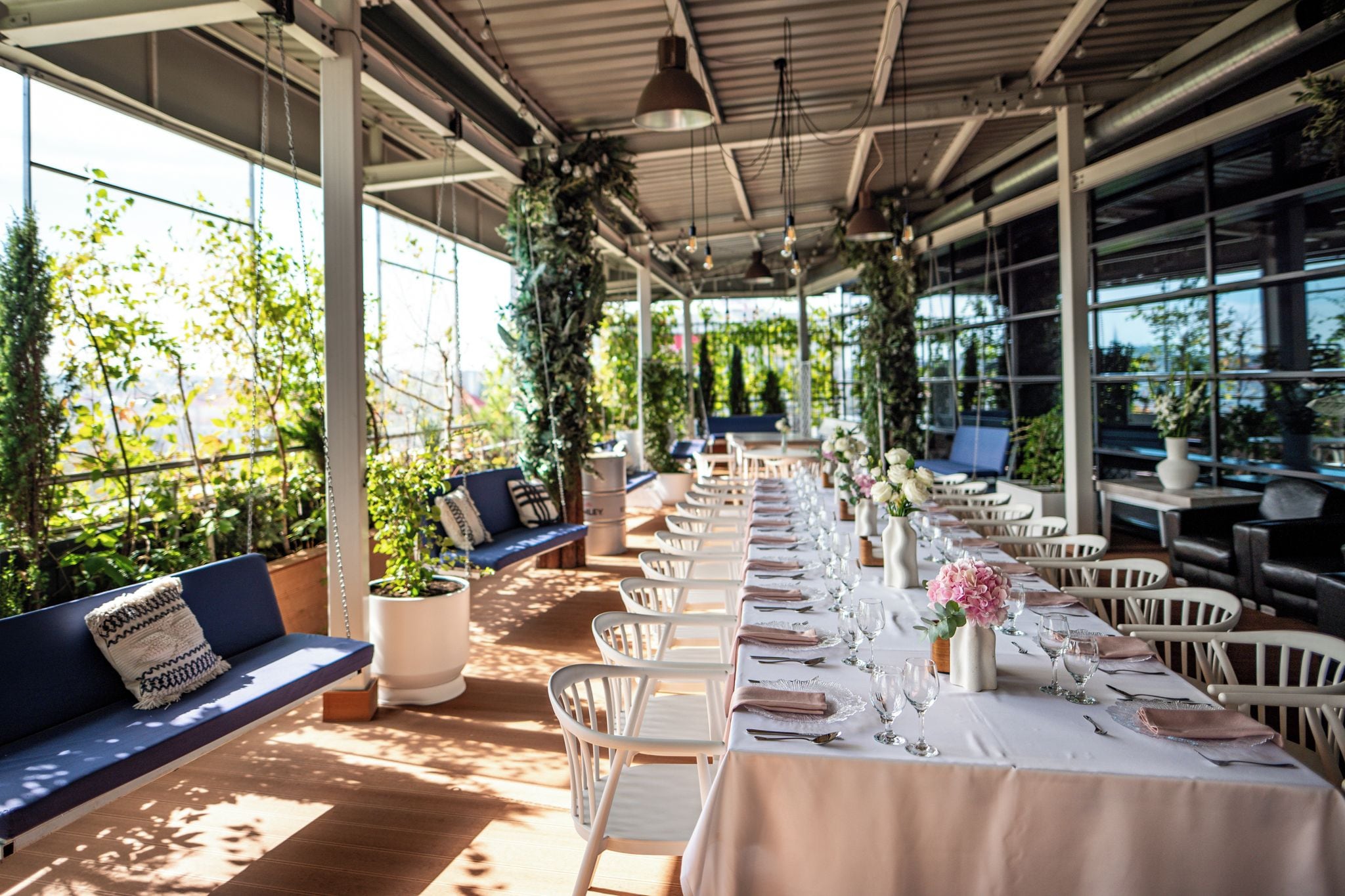 Un área de comedor al aire libre con mesas dispuestas para una comida, rodeada de sillas blancas y un largo banco con cojines.