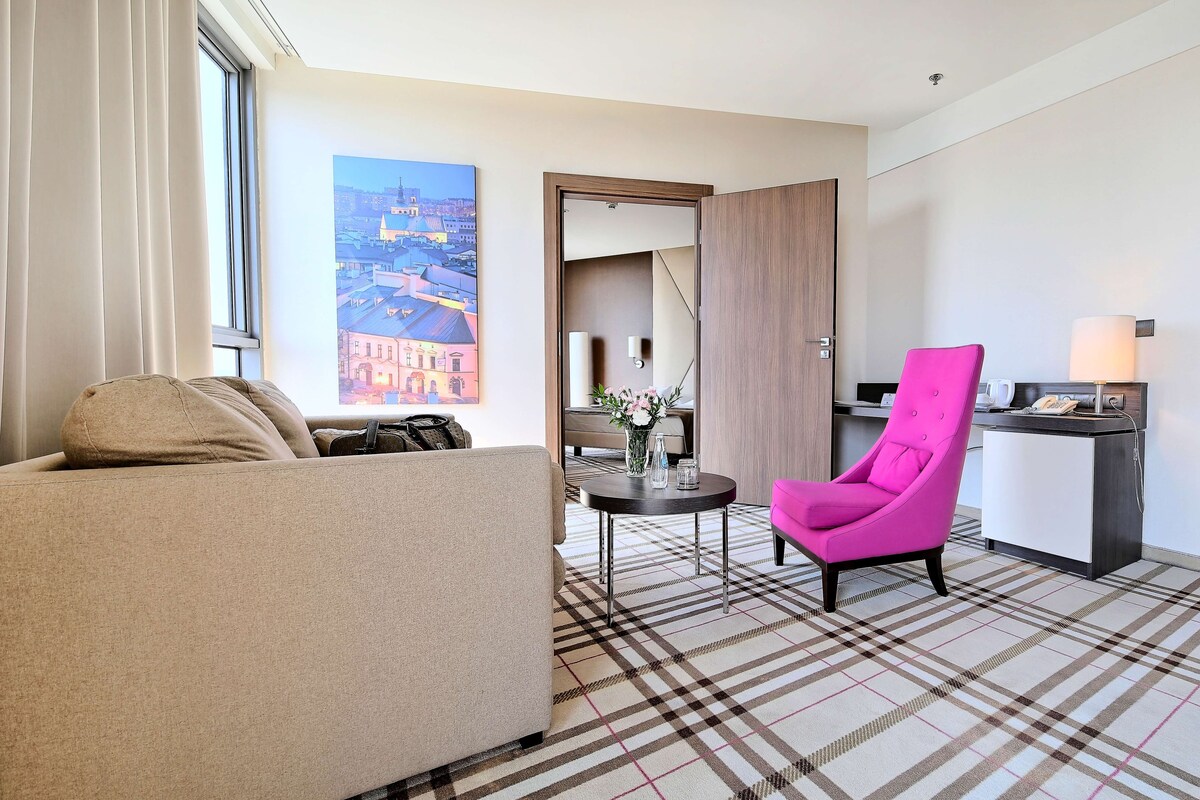 A hotel room with a sofa, a pink chair, a small table with a vase, and a view of buildings through a window.