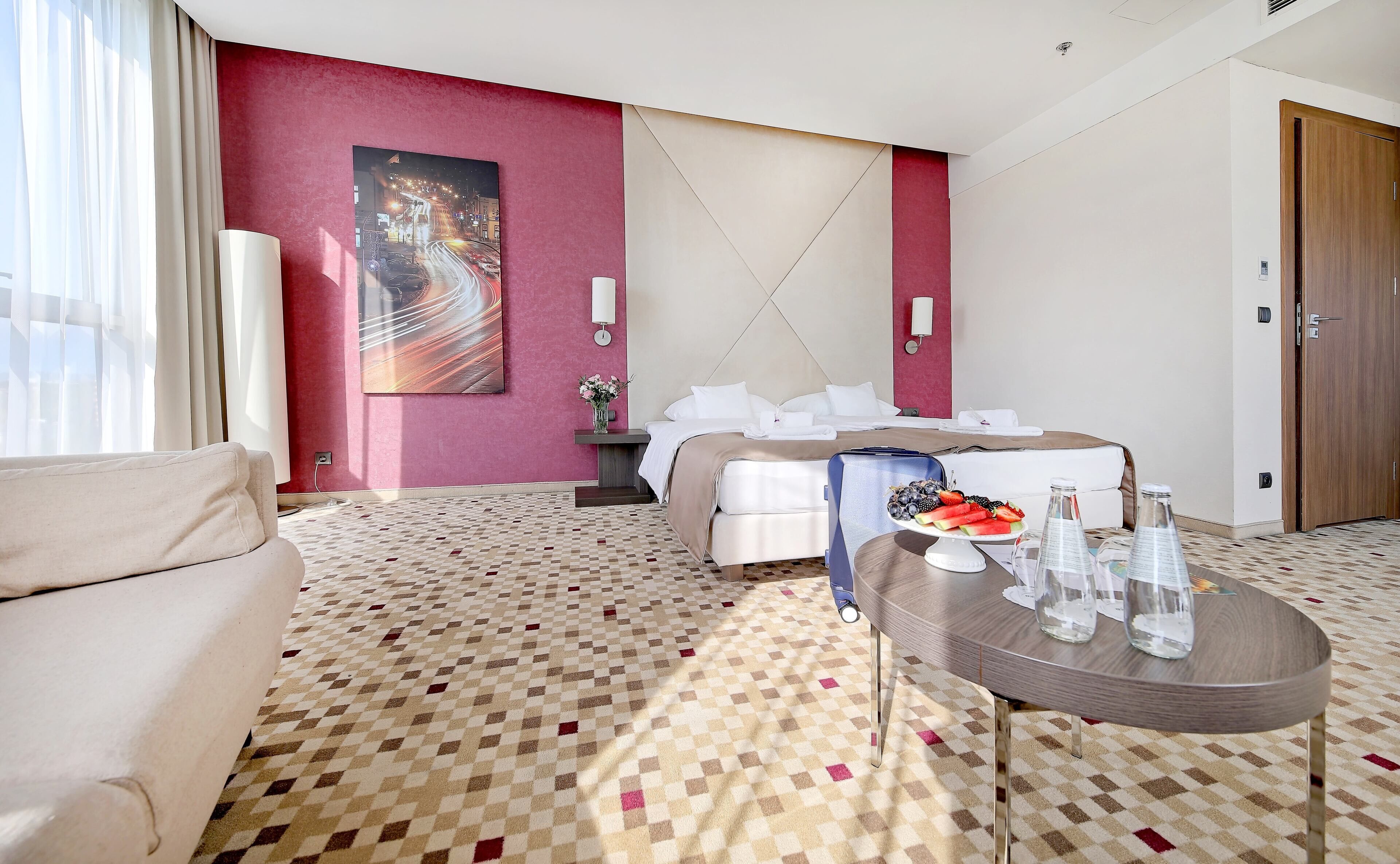 A hotel room with a bed, a coffee table with fruit and water bottles, a sofa, and a painting on the wall.