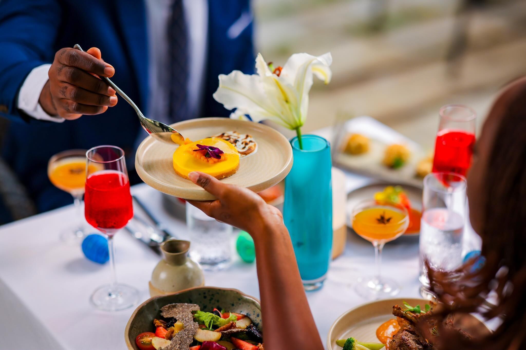 A dining table with various dishes and drinks, a vase with a white flower, and a person holding a plate.