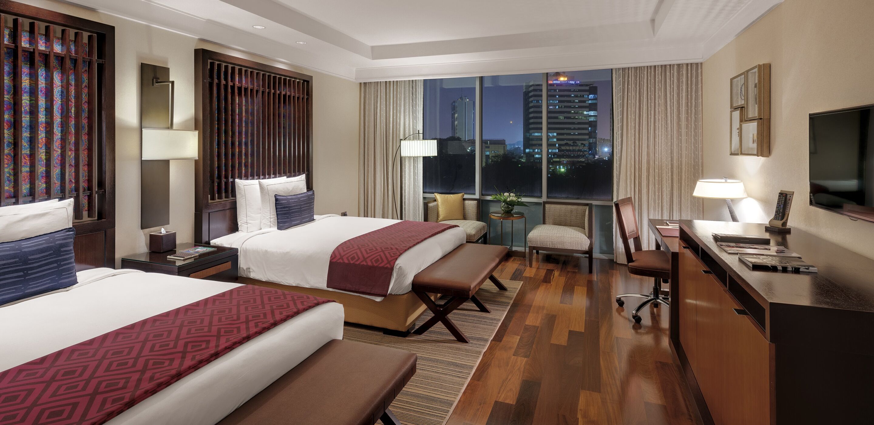 A hotel room with two beds, a desk, a chair, a TV, and a large window with a city view.