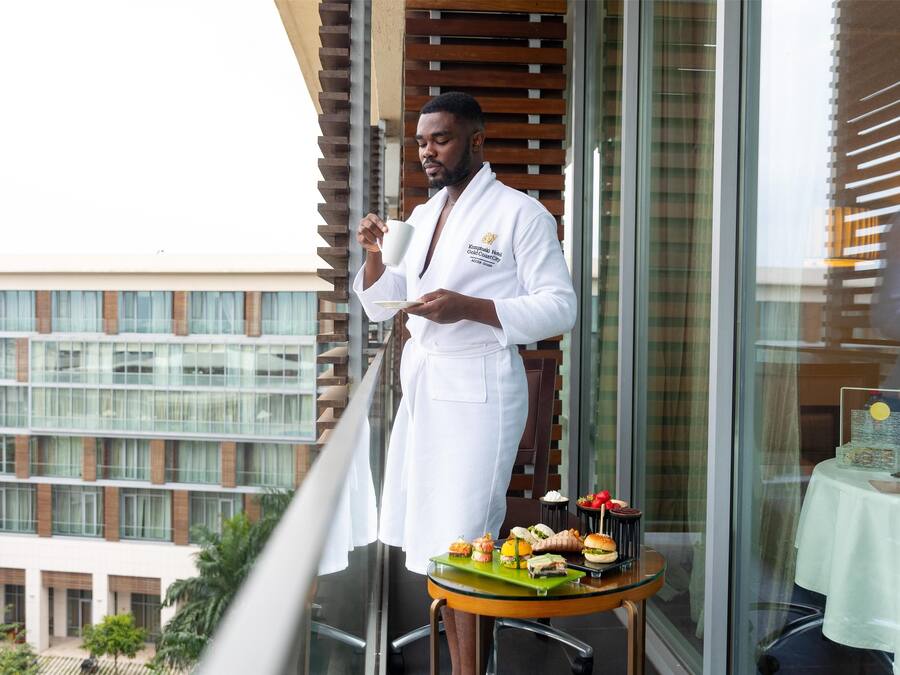 A person in a white robe standing on a balcony with a table of assorted snacks and a drink.