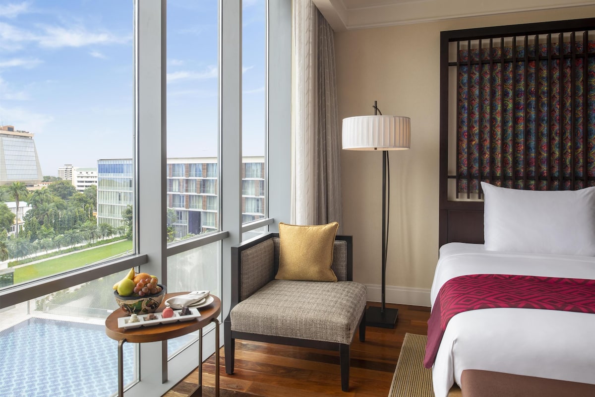 Ein Hotelzimmer mit großem Fenster, einem Bett, einem Sessel, einem Tisch mit Obst und einer Lampe.