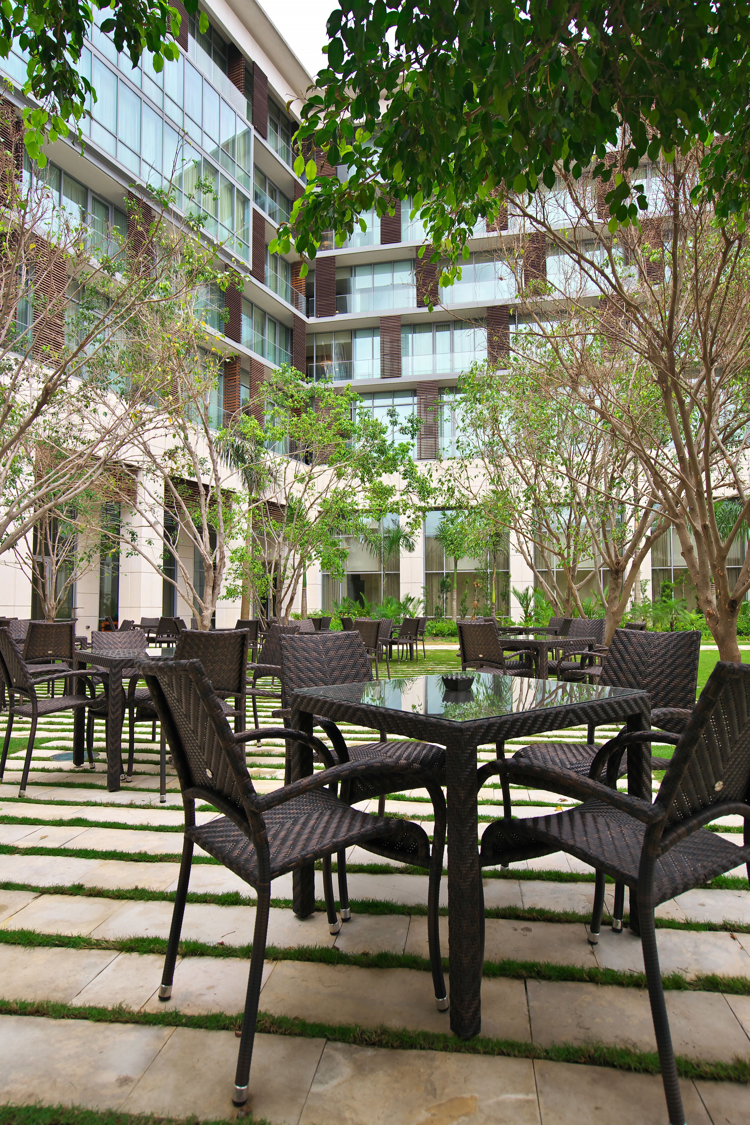An outdoor seating area with wicker furniture and a glass table, surrounded by greenery and a modern building in the background.