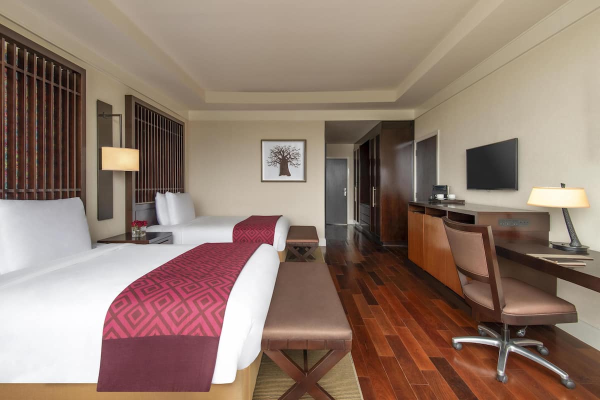 Ein Hotelzimmer mit zwei Betten, einem Schreibtisch, einem Sessel, einem Fernseher und einem im Hintergrund sichtbaren Badezimmer.