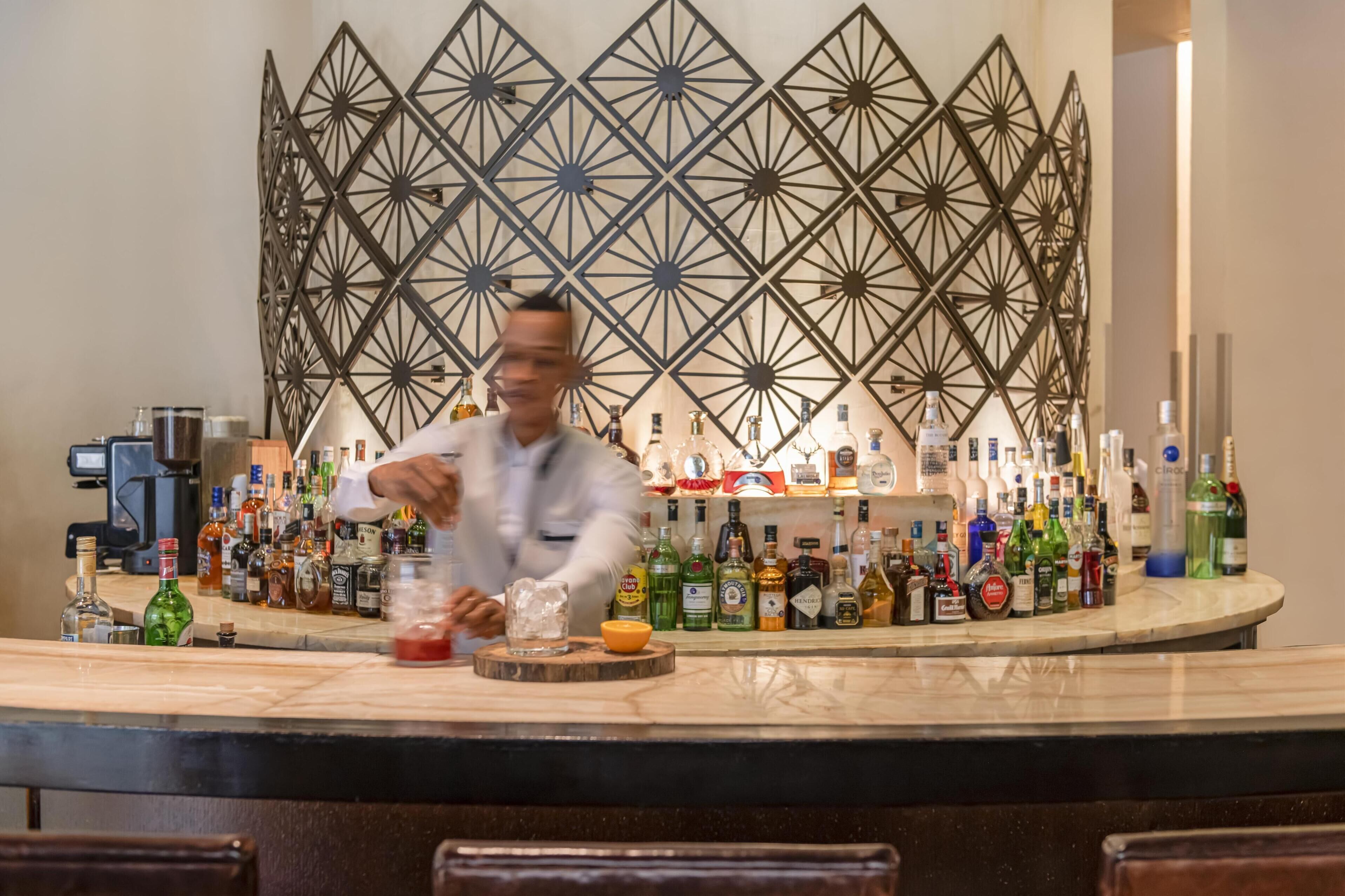 A bar area with a bartender preparing drinks, a variety of liquor bottles, and a decorative geometric wall in the background.