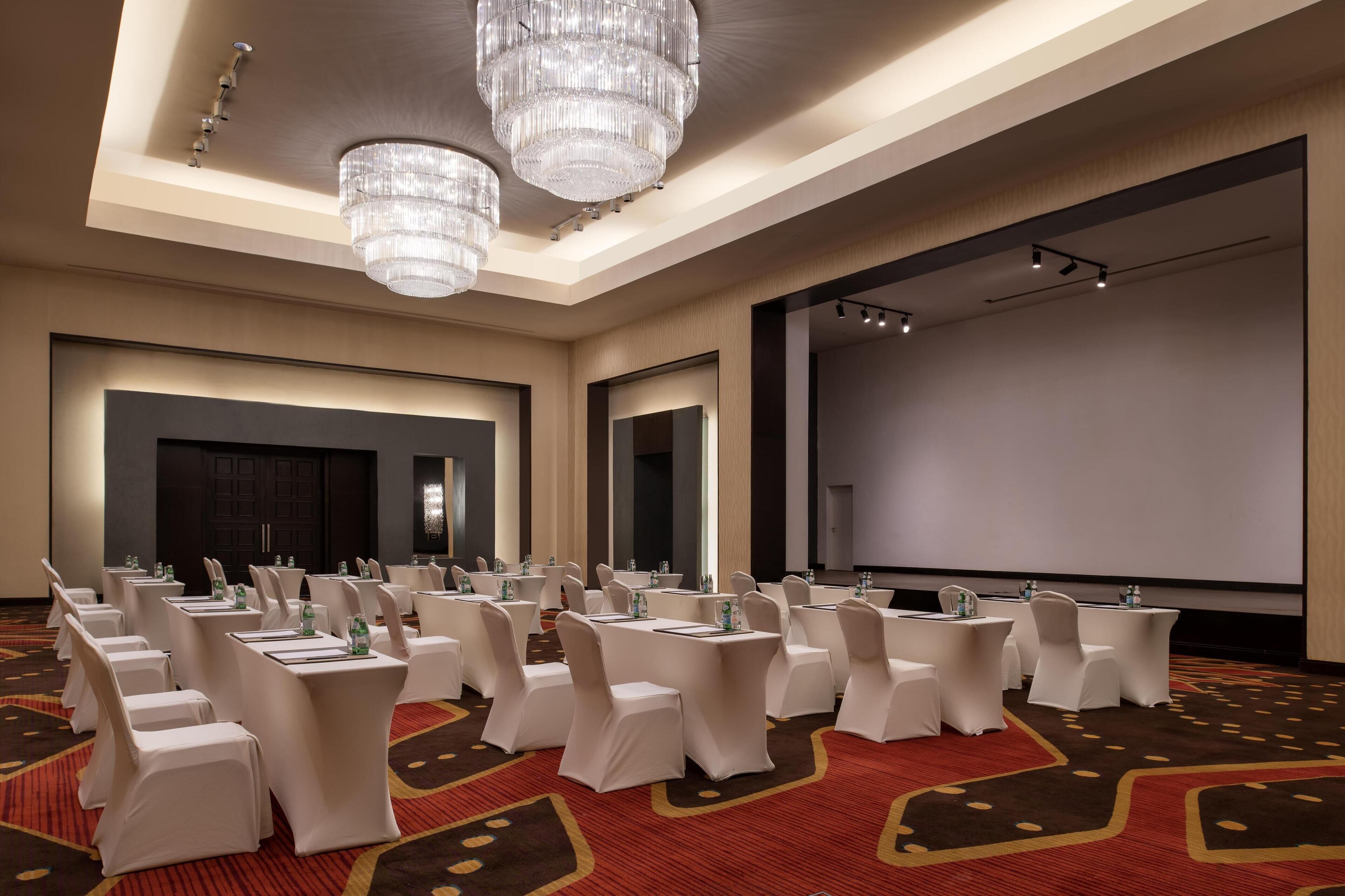 Un salón de conferencias con filas de mesas y sillas blancas, una pantalla grande y candelabros.
