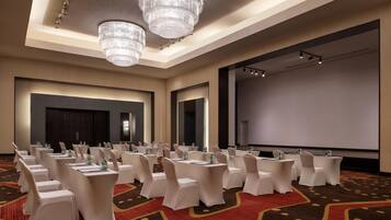 Un salón de conferencias con filas de mesas y sillas blancas, una pantalla grande y candelabros.