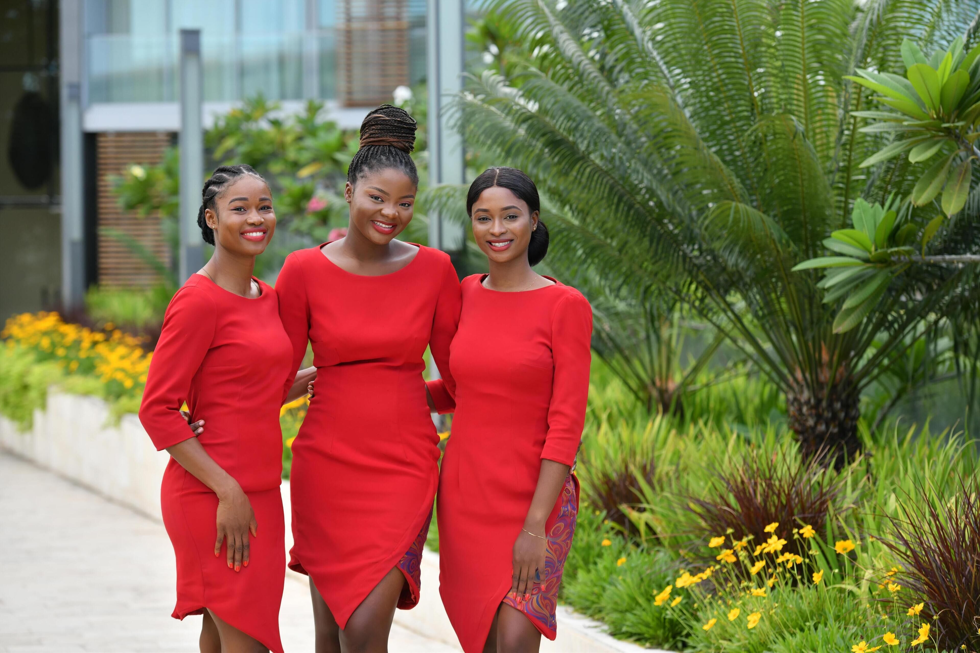 Three women in red dresses walking on a pathway surrounded by greenery and yellow flowers.