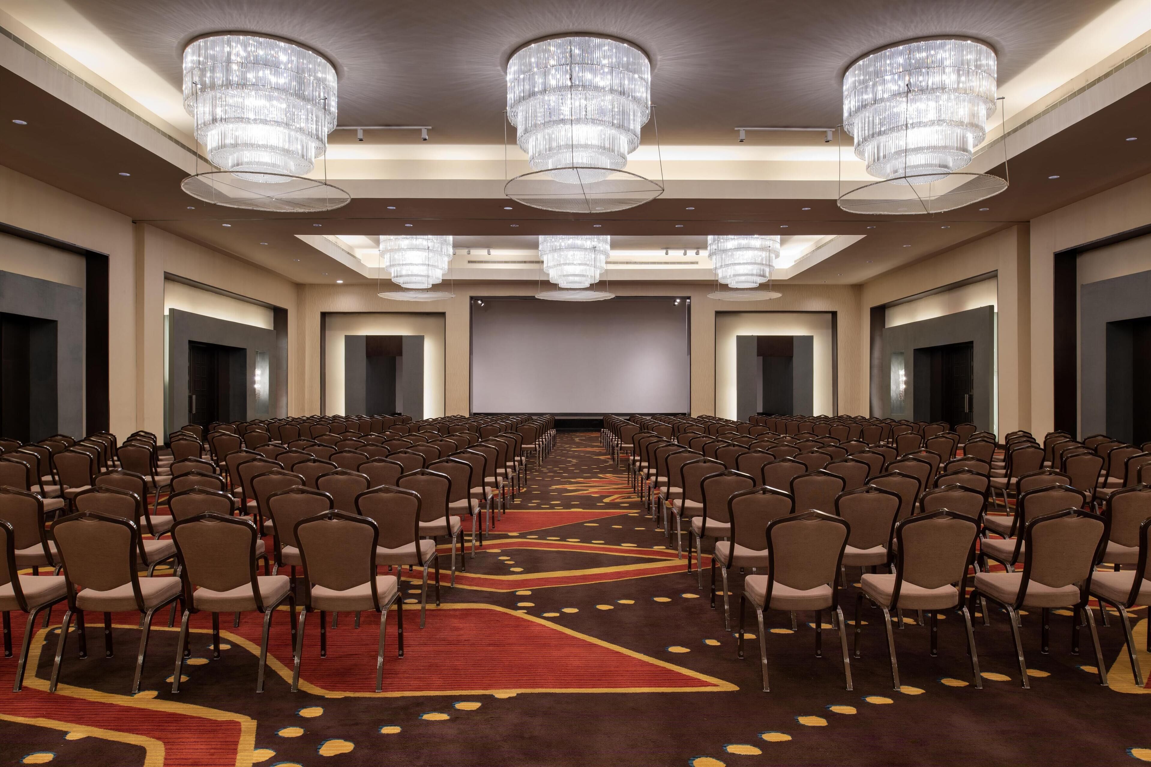 Un salón de conferencias con filas de sillas, una pantalla grande y candelabros.