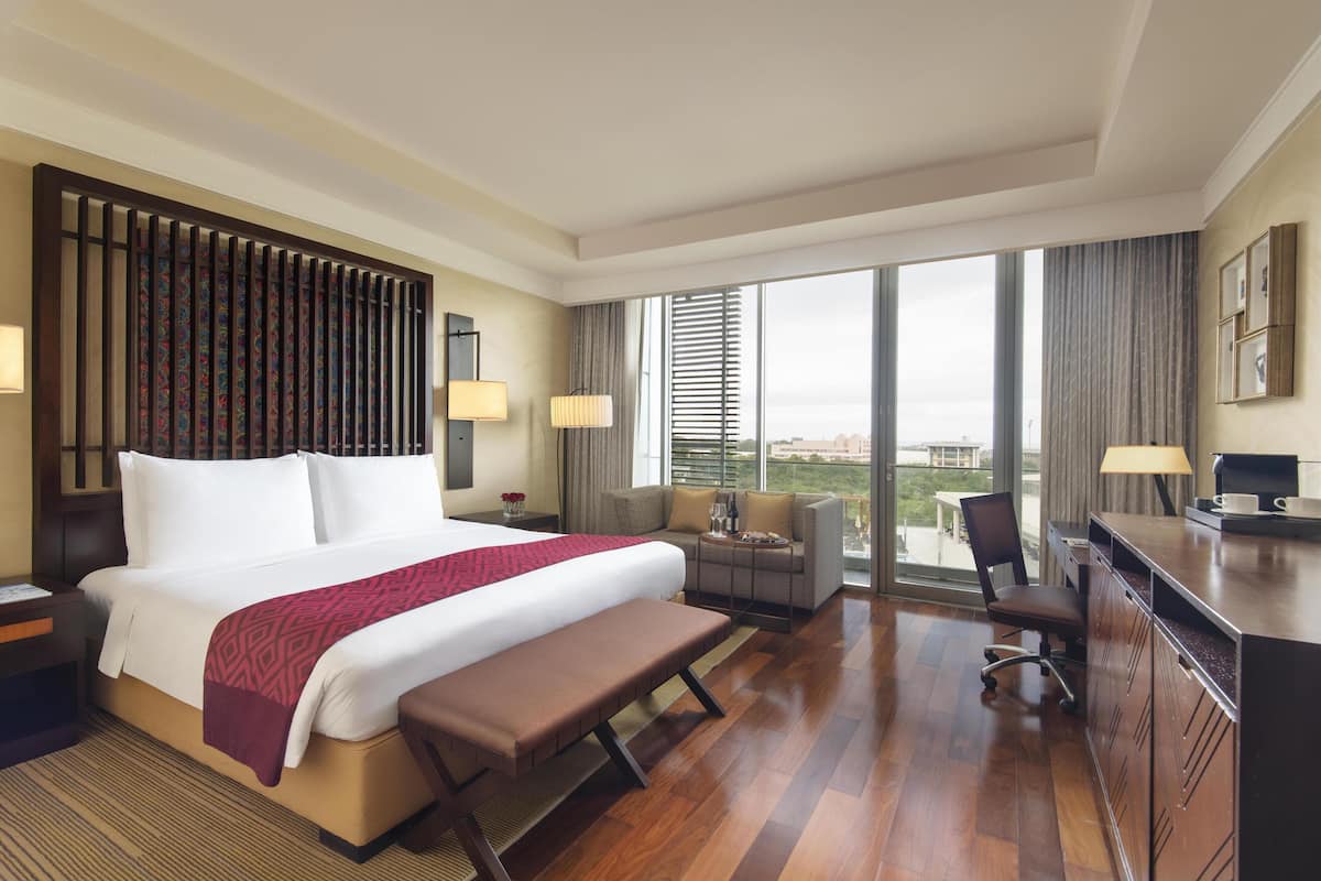 Ein Hotelzimmer mit einem großen Bett, einem Schreibtisch, einem Stuhl und einer Couch. Ein großes Fenster bietet Blick auf die Stadt.