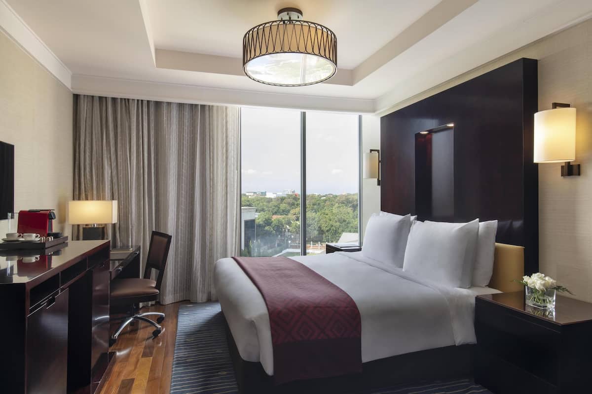 Ein Hotelzimmer mit einem großen Bett, einem Schreibtisch mit Lampe, einem Sessel und Blick auf die Stadt durch große Fenster.