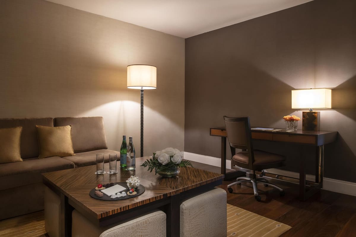 Ein Hotelzimmer mit einer Couch, einem Esstisch mit Blumenvase, zwei Lampen und einem Schreibtisch mit Stuhl.