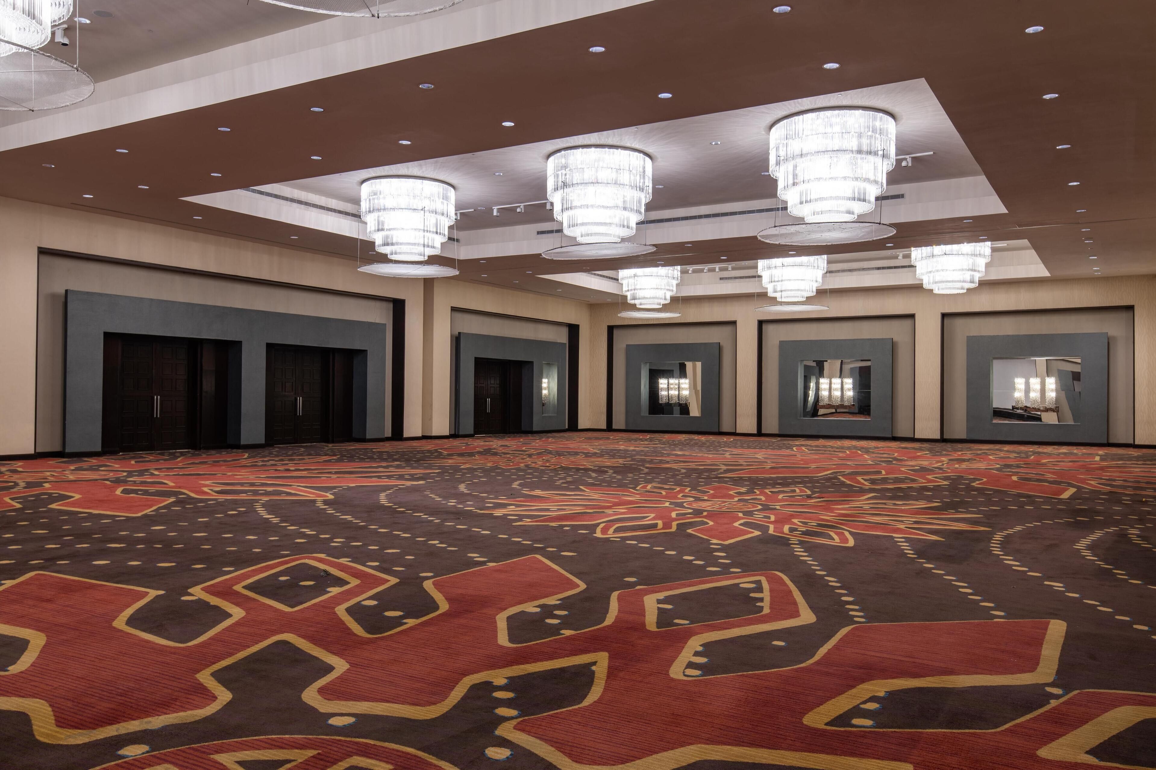 Un amplio salón de conferencias con alfombra estampada, varias puertas y grandes candelabros.