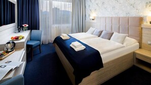 A hotel room with a large bed, a desk with a chair, a small table with a kettle and fruit, and a window with curtains. - Park Hotel (Dolny Kubin)