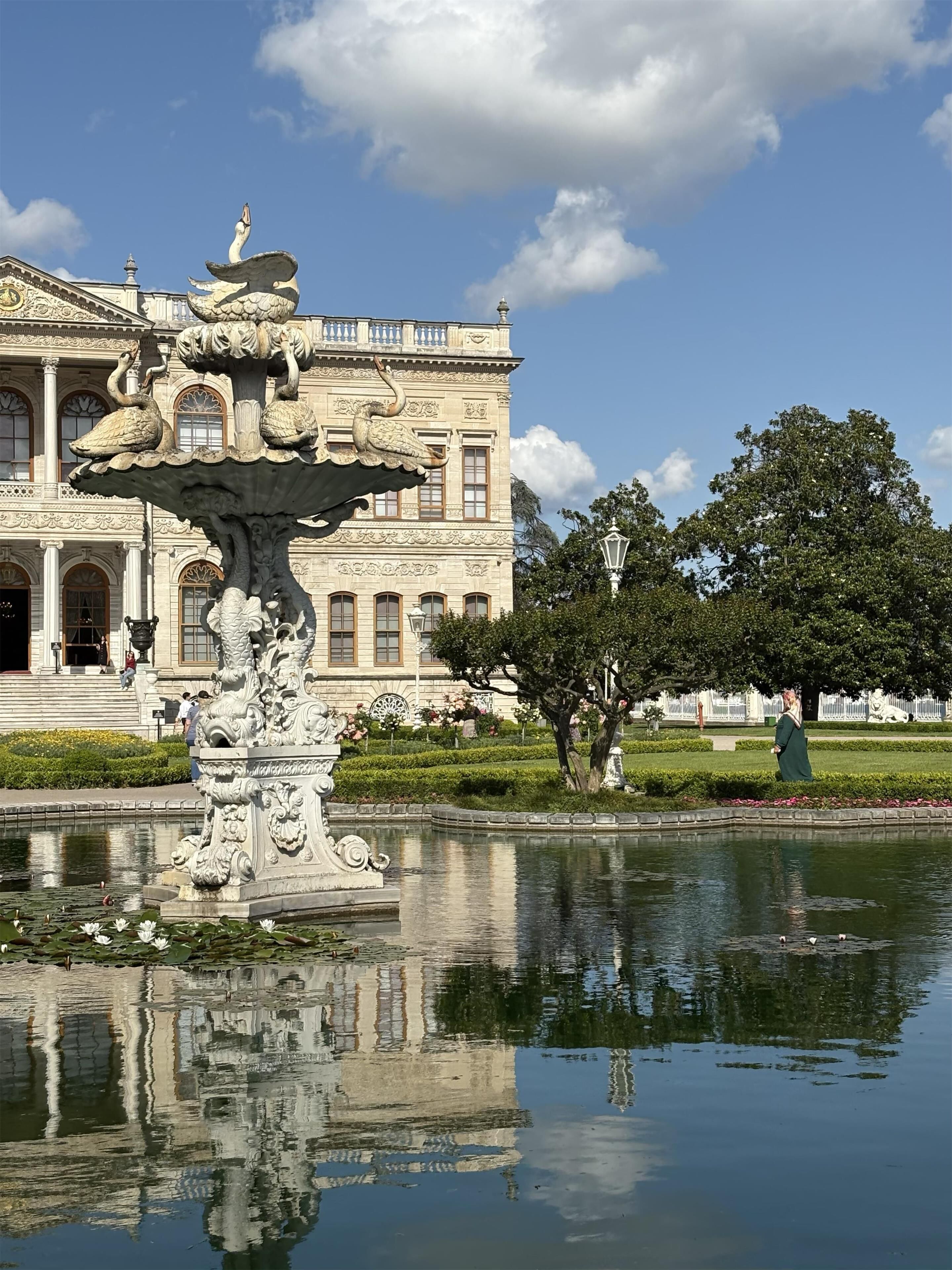 A classical fountain with ornate sculptures in front of a grand building with arched windows and a symmetrical facade.