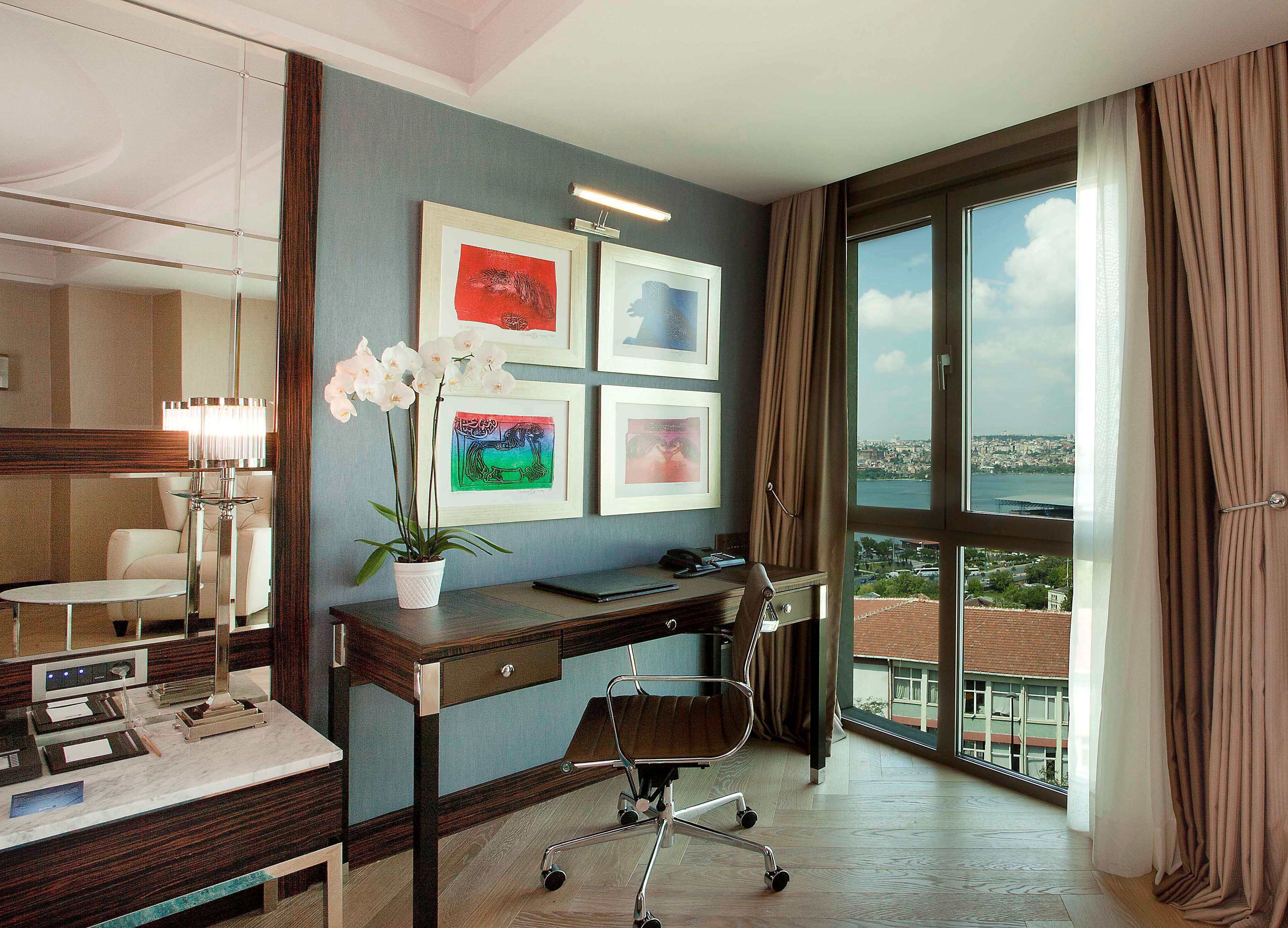 A hotel room with a desk, chair, and a potted orchid. There are framed artworks on the wall and a view of the city through the window.