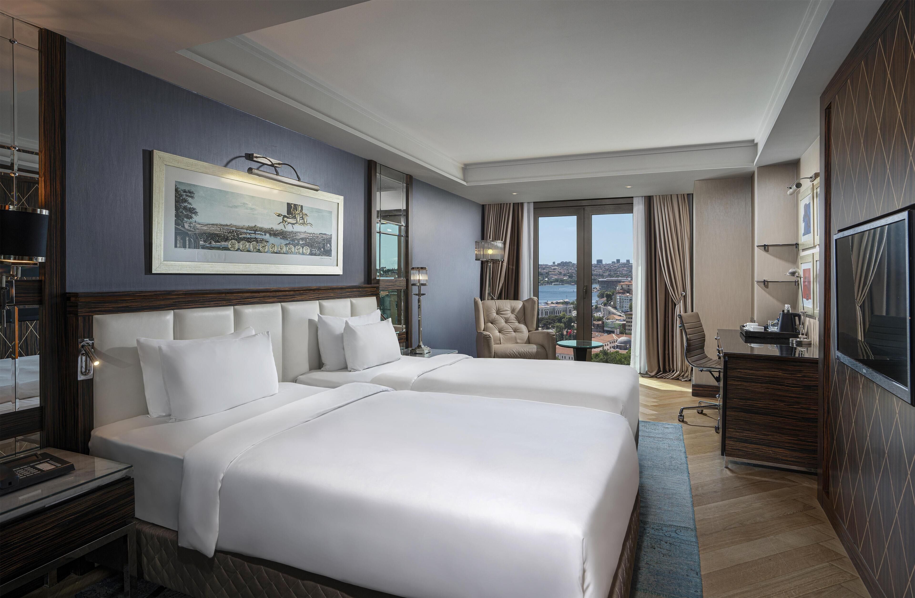 A hotel room with two large beds, a desk, a chair, a TV, and a large window with a city view.