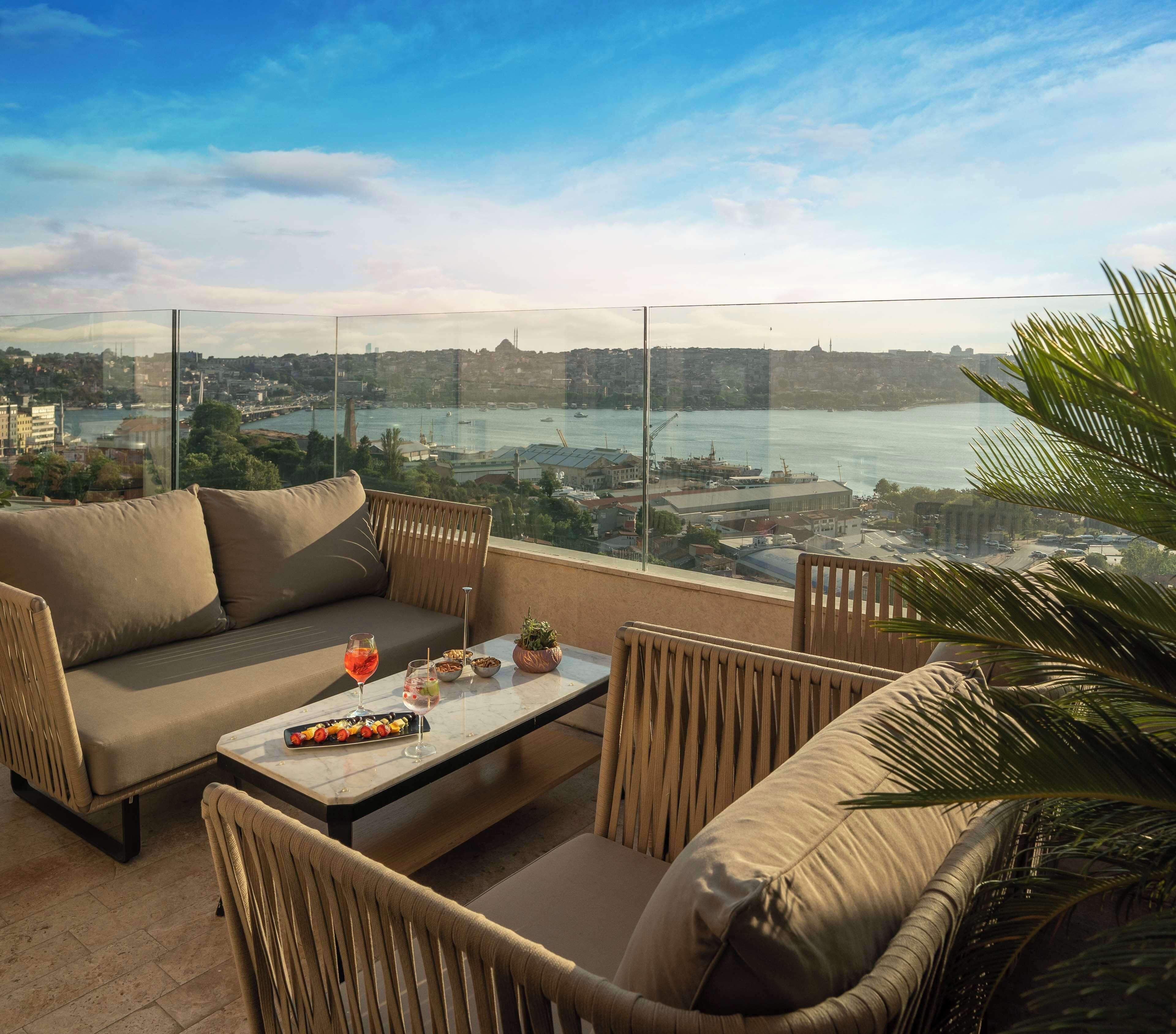 A balcony with a sofa, chairs, and a table with food and drinks. A cityscape and body of water are visible in the background.