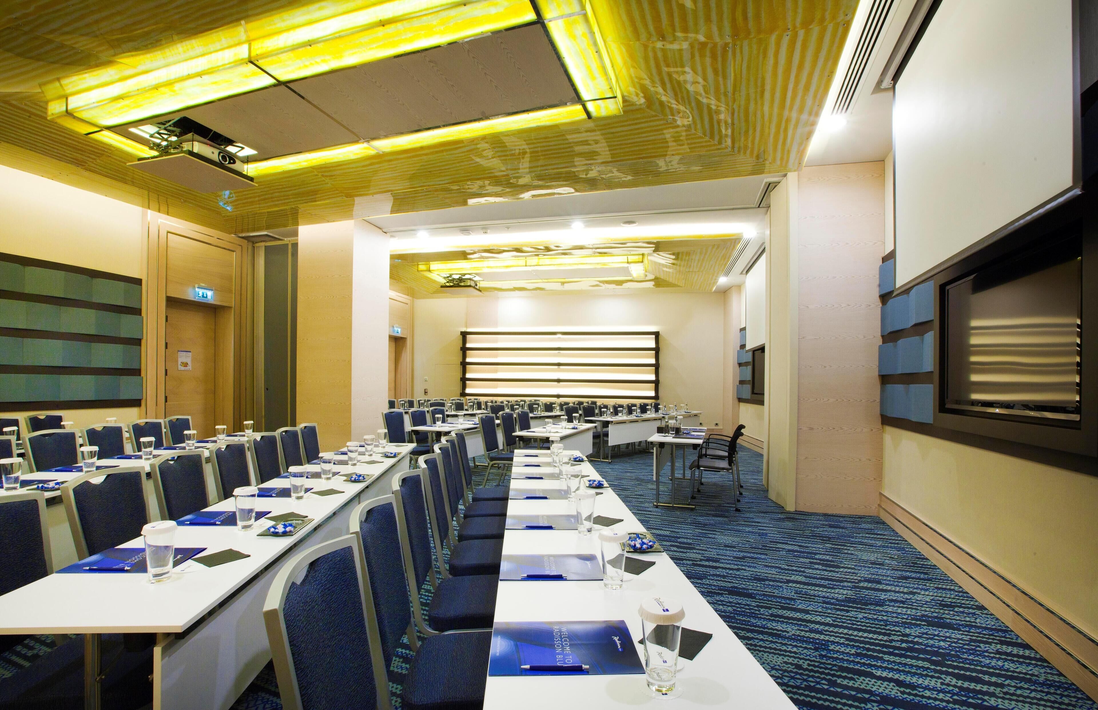 A conference room with rows of tables and chairs, a projector, and a large screen.