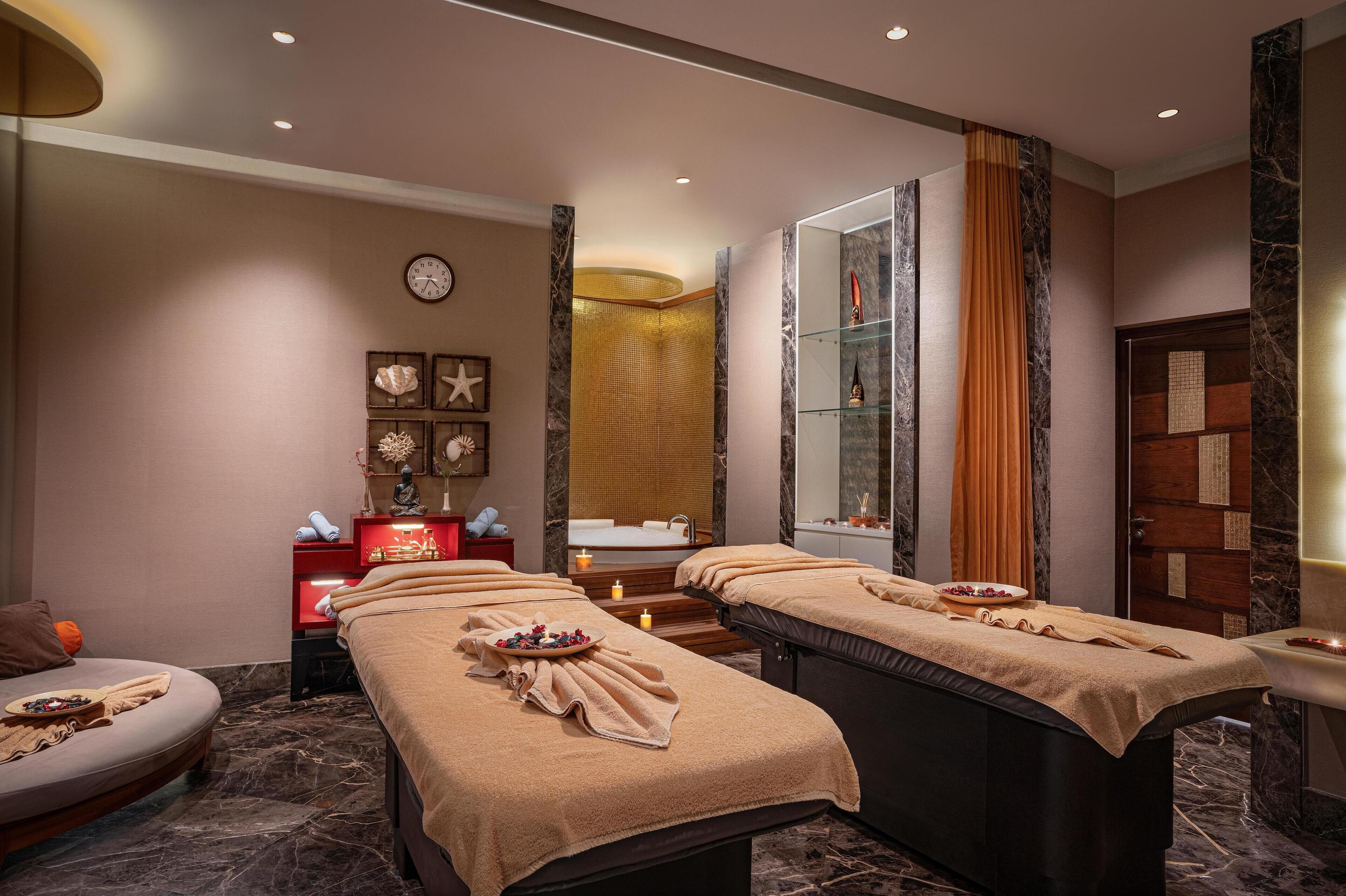 Ein Spa-Behandlungsraum mit zwei Massagetischen, einer Sitzecke, einem Spiegel und einem kleinen Waschbecken.