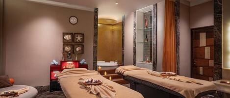 A spa room with two massage tables, a small seating area, a mirror, and a small sink.