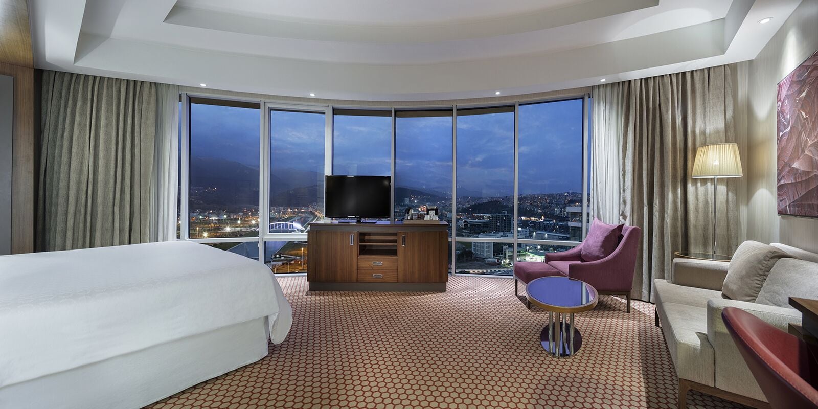 Ett hotellrum med en stor säng, en TV, ett skrivbord och utsikt över en stadssiluett i nattljus.